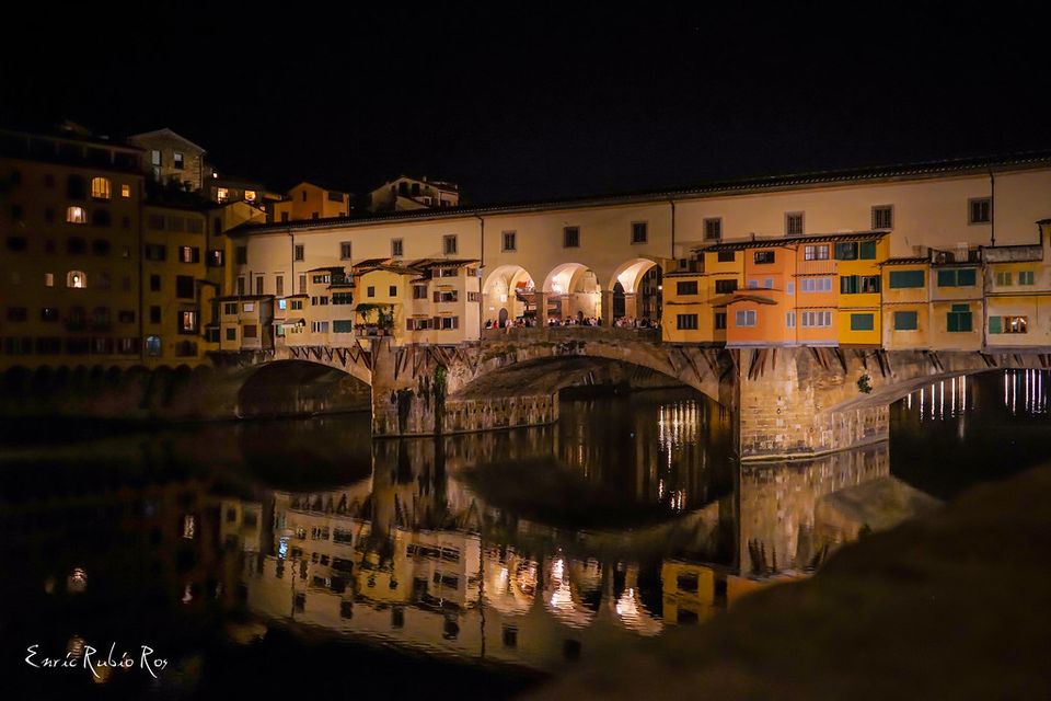 Il Ponte Vecchio di notte, botteghe e arcate illuminate riflesse nel fiume
